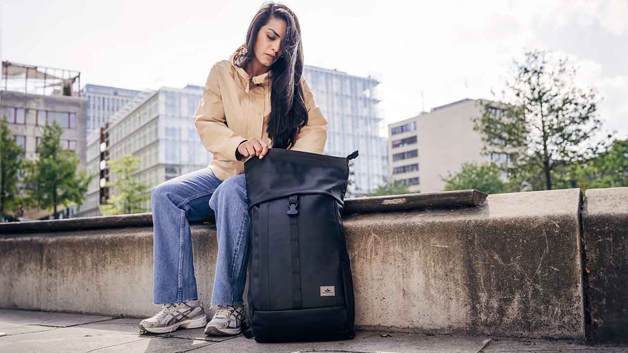 Eine Person sitzt auf einer Mauer und hält einen schwarzen Rucksack von Freibeutler. Sie trägt eine beige Jacke und blaue Jeans.