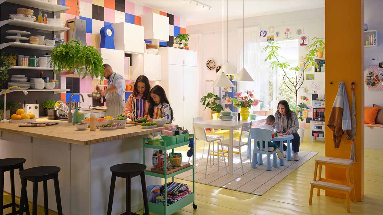 Helle Wohnküche mit bunter Wandgestaltung, Kücheninsel, mehreren Personen, Pflanzen und Kinderspielecke mit kleinem Tisch und Stühlen