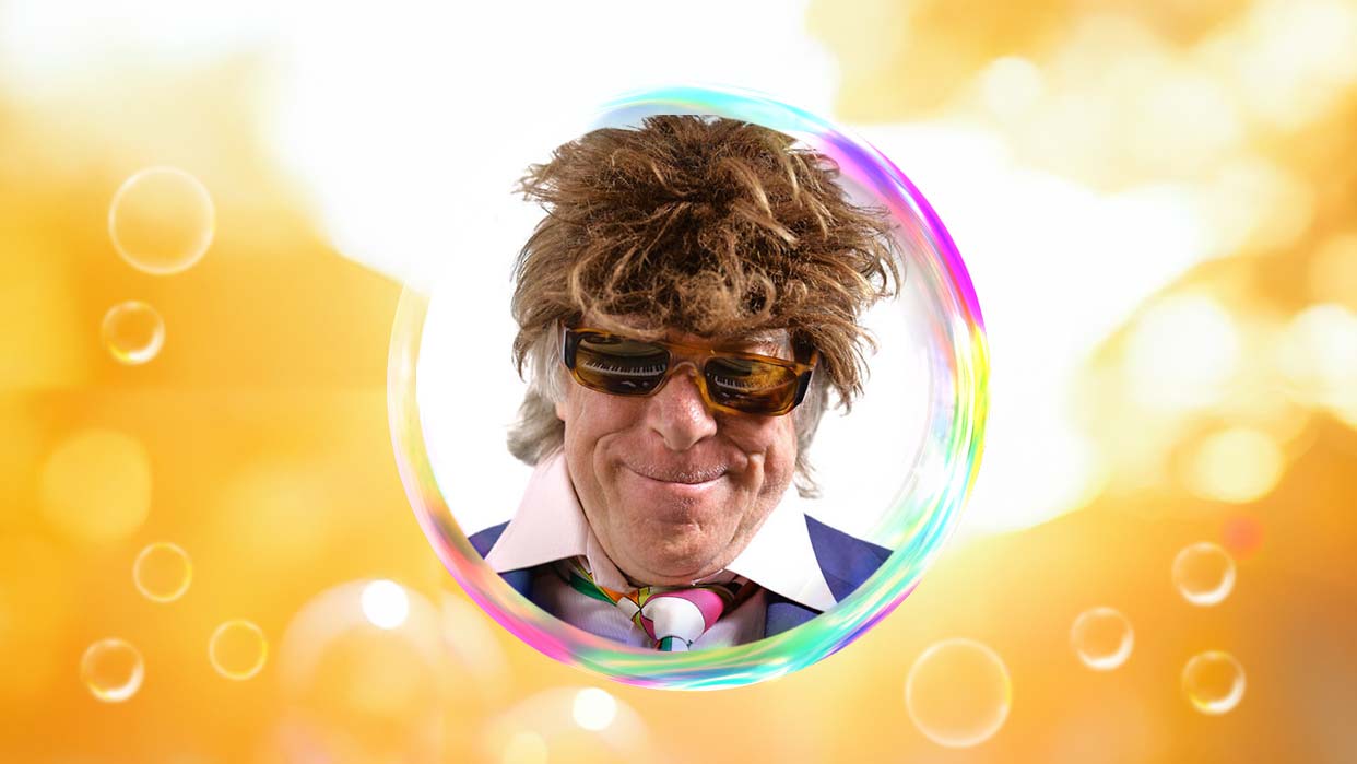 Helge Schneider mit brauner Perücke, Sonnenbrille und bunter Krawatte, umgeben von einer großen Seifenblase vor gelbem Hintergrund.