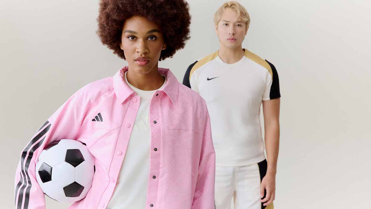 Zwei Personen stehen nebeneinander, eine trägt ein rosa Hemd und hält einen Fußball, die andere ein weißes Sportshirt mit schwarzen und goldenen Ärmeln.