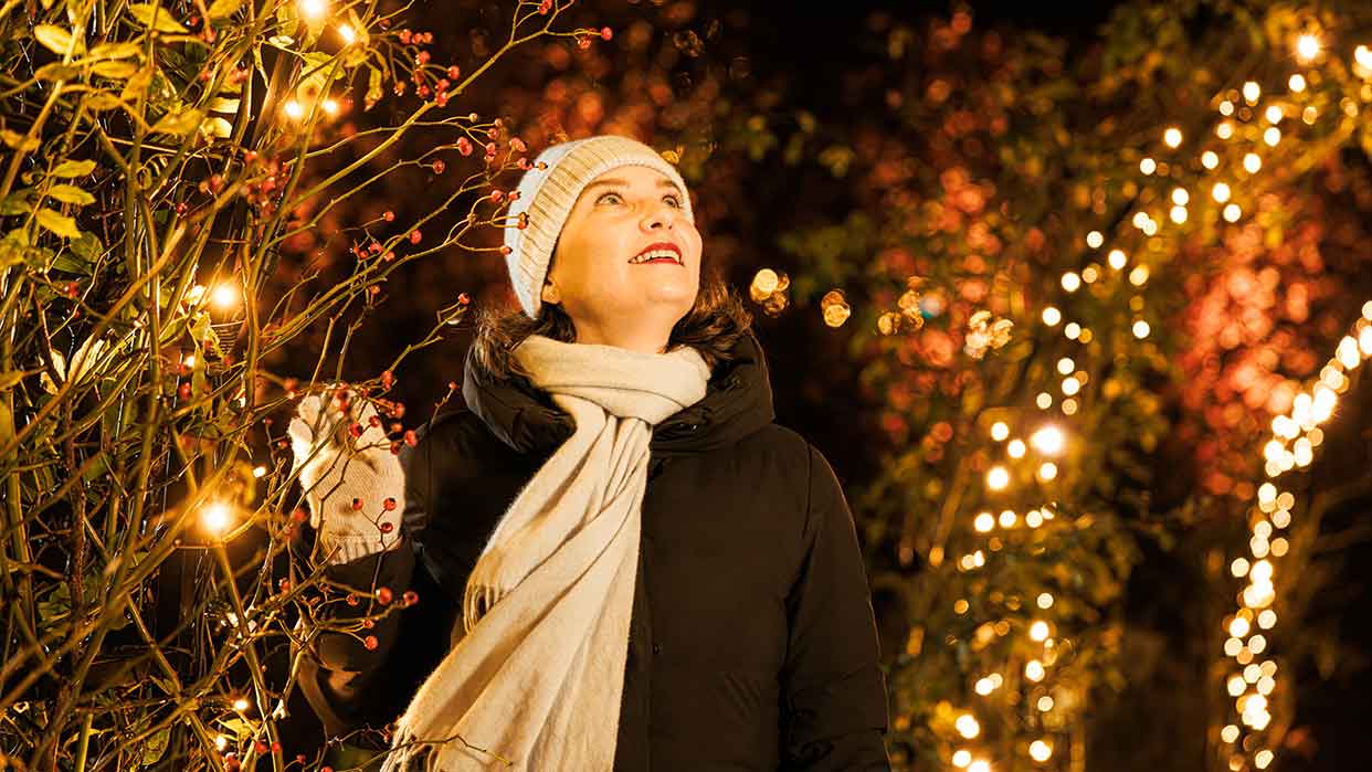 Person in schwarzer Winterjacke, weißem Schal und weißer Mütze steht in einem Garten mit warmen Lichterketten und roten Beeren an den Ästen.