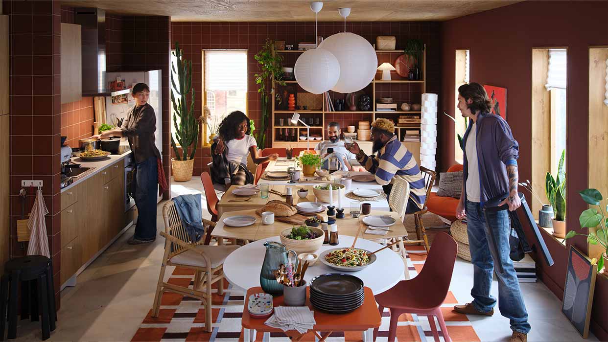 Esszimmer mit langem Tisch, gedeckt mit Tellern und Speisen, umgeben von mehreren Personen in einem Raum mit braunen Fliesen und großen Fenstern.