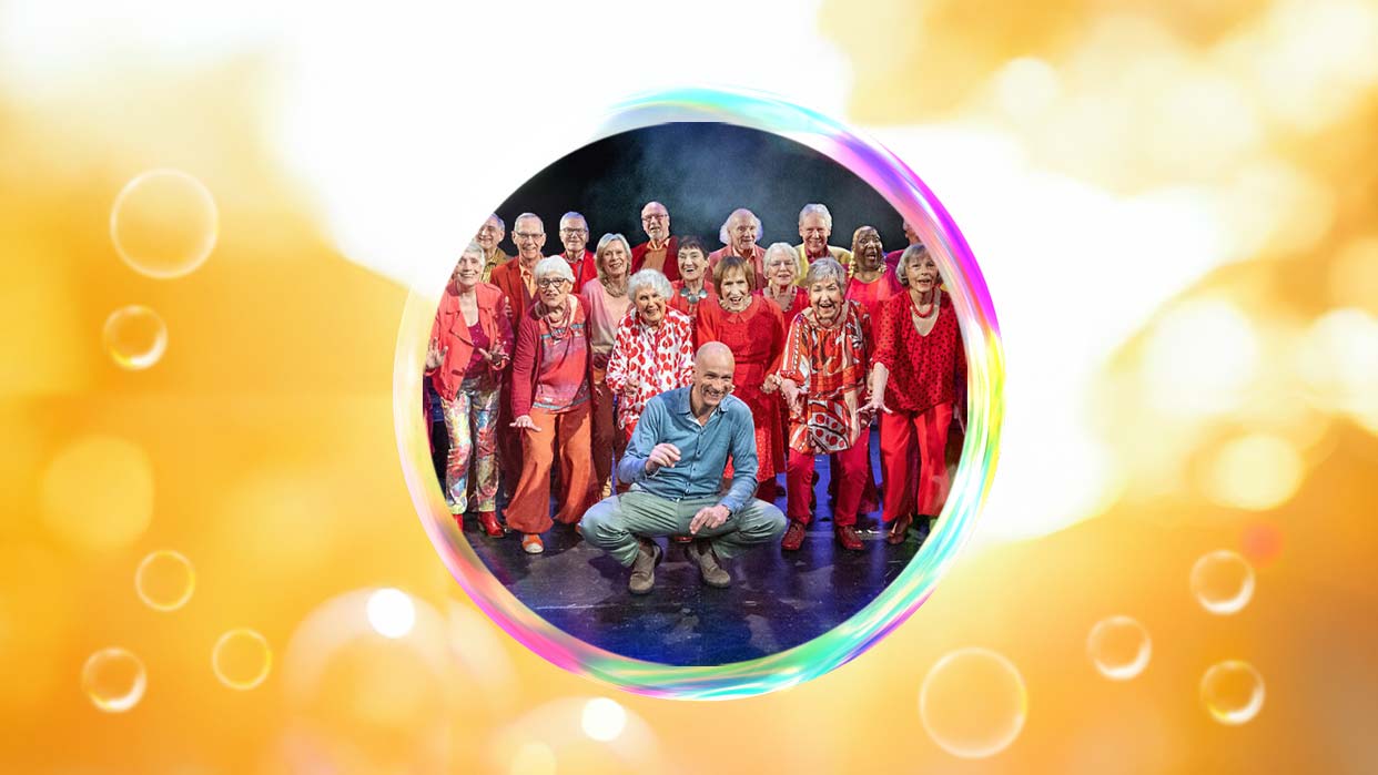 Heaven Can Wait Chor, die in bunten roten Outfits auf einer Bühne stehen, vor einem gelb-orangen Hintergrund mit Seifenblasenmotiv.