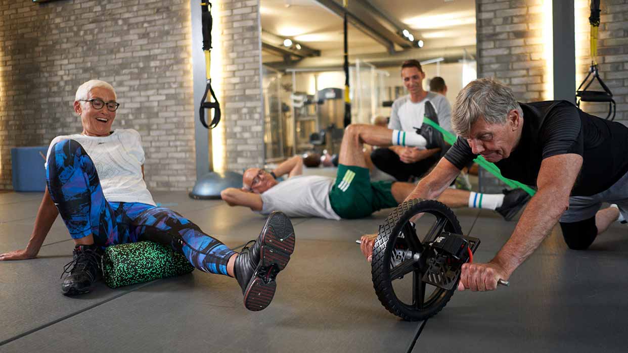 Mehrere Personen trainieren in einem Fitnessstudio, eine Person nutzt eine Faszienrolle, eine andere ein Bauchmuskelrad, weitere dehnen sich auf der Matte.
