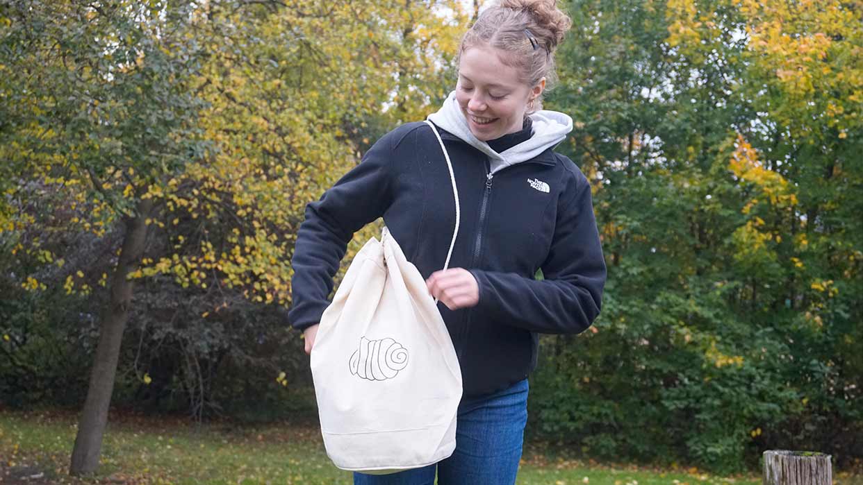 Frau trägt schwarzen Fleecepullover und hält einen hellen, runden Seesack mit Kordelzug und Franzbrötchen Muster vor einem herbstlichen Wald.
