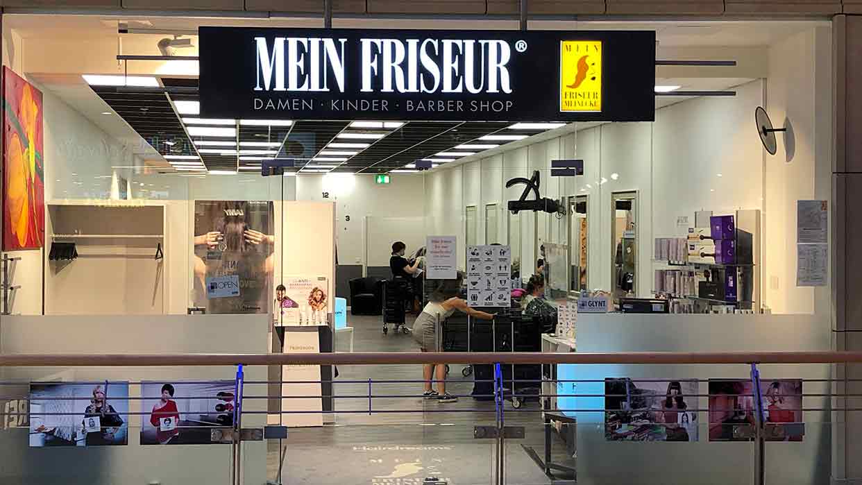Innenansicht des Friseursalons ‚Mein Friseur Meinecke‘ mit Friseuren und Kunden bei der Arbeit; moderner Salon für Damen, Herren und Kinder.
