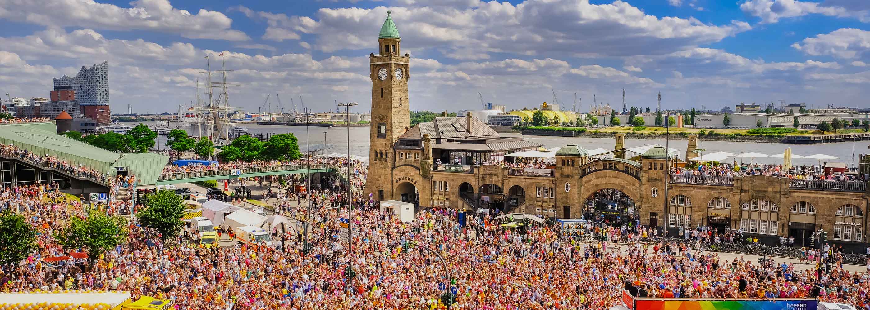 Menschenmenge vor dem historischen Landungsbrücken-Gebäude mit Turm an der Elbe in Hamburg bei sonnigem Wetter.