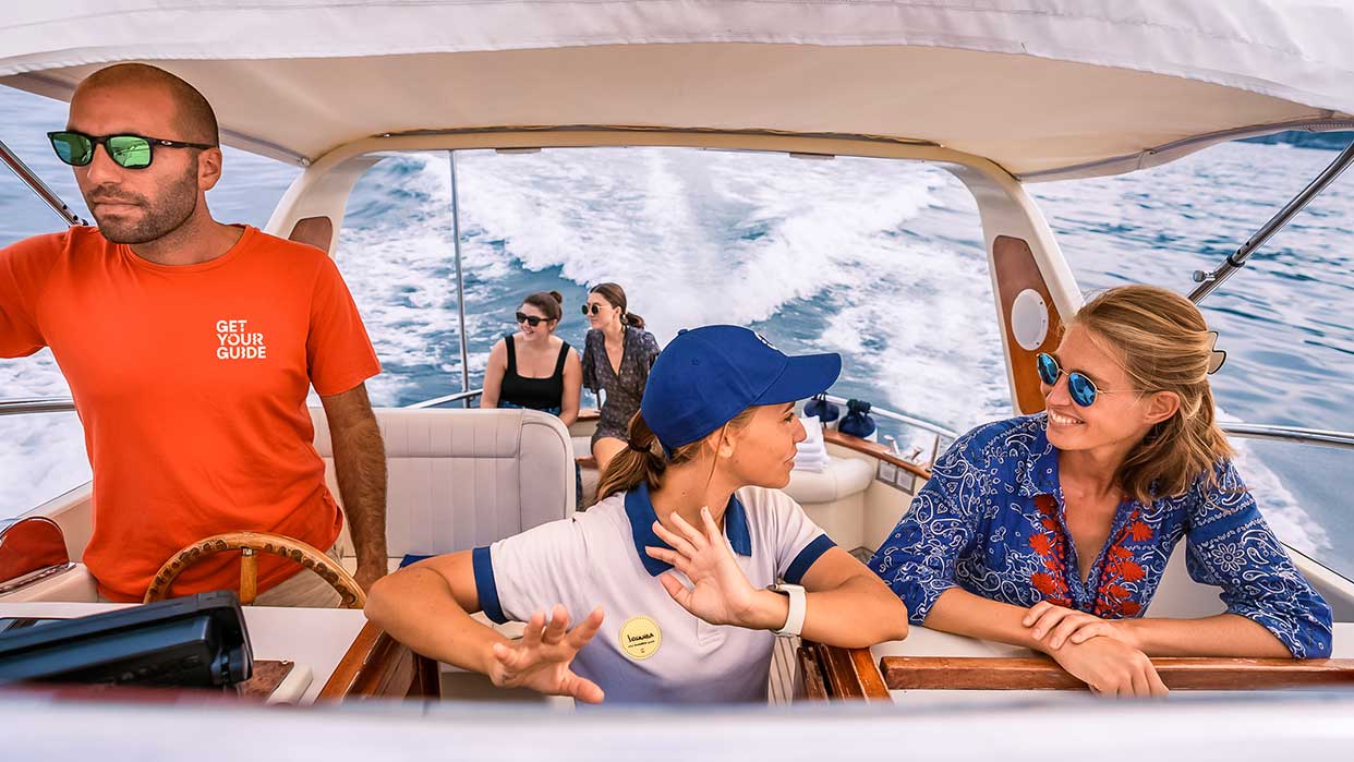 Ein GetYourGuide-Mitarbeiter in einem roten T-Shirt steuert ein Boot, auf dem sich mehrere Personen befinden. Das Boot fährt über ruhiges Wasser im Rahmen einer geführten Tour.