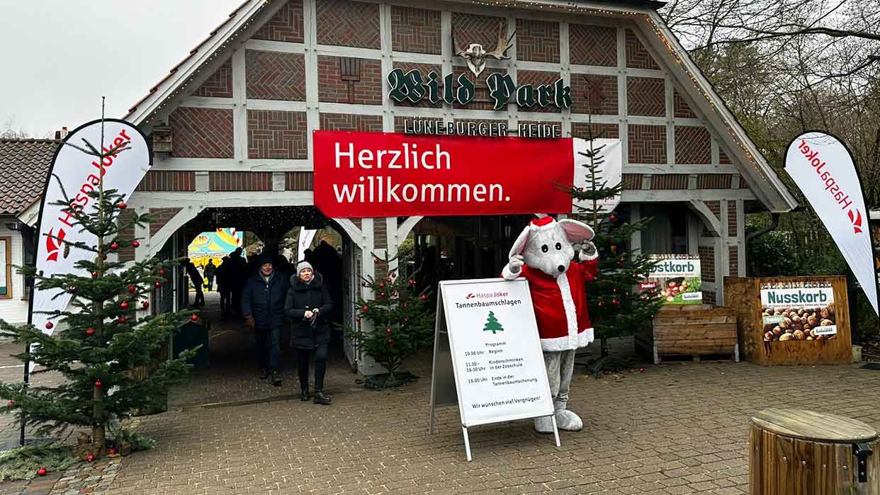 Eingang zum Wildpark mit Fachwerkhaus, zwei Tannenbäumen, rotem Banner mit 'Herzlich willkommen.' und Person im Weihnachtsmaus-Kostüm neben HaspaJoker Schild.