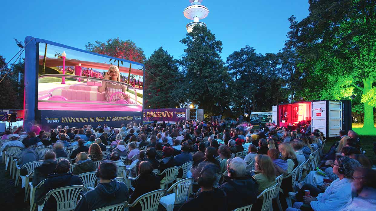 Publikum sitzt bei Nacht im Open-Air-SchanzenKino Hamburg und schaut einen Film auf großer Leinwand mit einer Frau am Steuer eines pinken Autos.