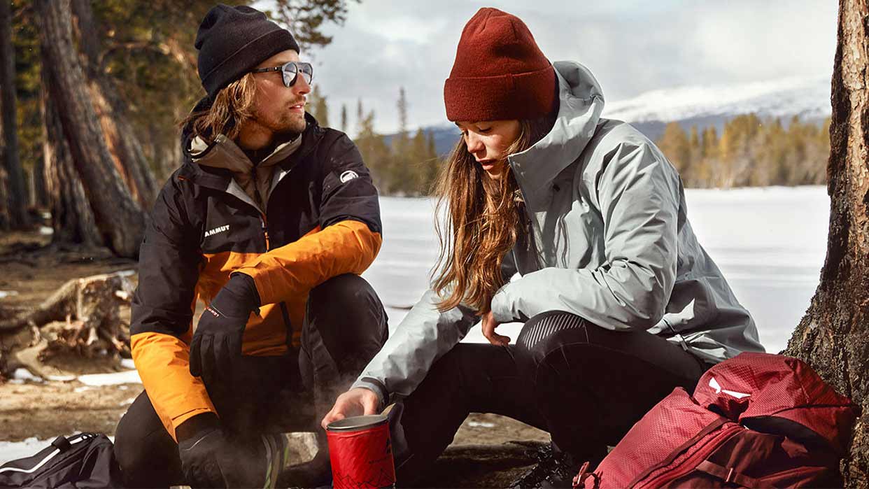 Zwei Personen in Outdoor-Bekleidung sitzen an einem See, eine hält einen roten Becher, im Hintergrund Bäume und Berge.