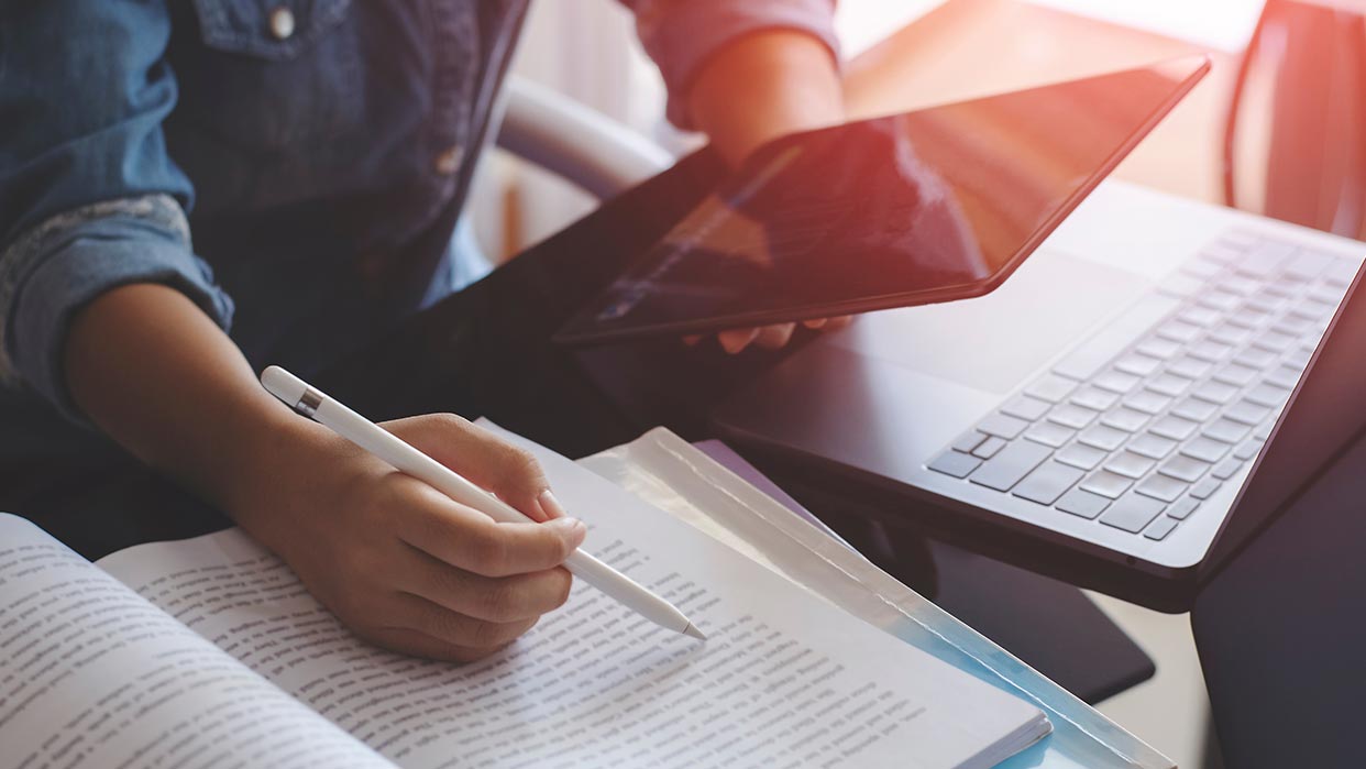 Person hält einen Stift und macht Notizen in einem aufgeschlagenen Buch, daneben liegt ein Laptop und eine weitere Person hält ein Tablet.