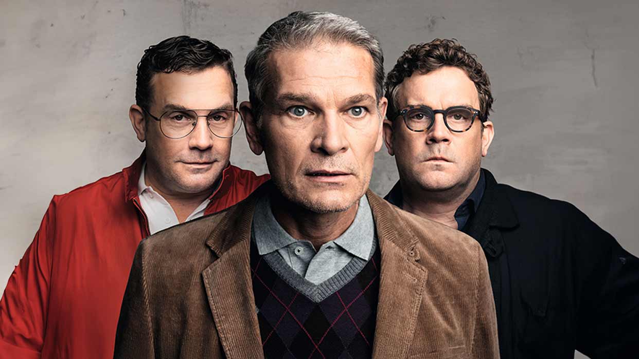 Drei Männer stehen nebeneinander, der mittlere trägt eine braune Jacke und ein kariertes Hemd, die anderen tragen rote beziehungsweise schwarze Oberteile.