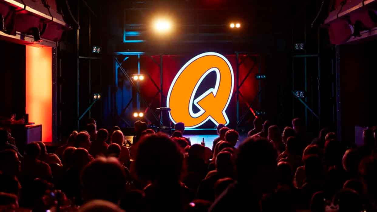 Große orangefarbene Buchstaben Q auf einer Bühne vor einem Publikum in einem dunklen Raum mit Bühnenbeleuchtung.