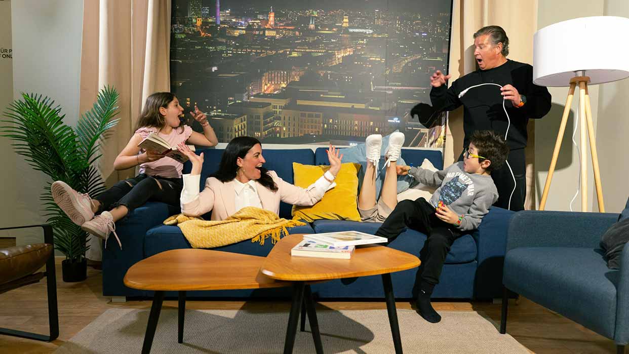 Vier Personen sitzen und stehen in einem Wohnzimmer mit blauer Couch, gelben Kissen, großem Stadtbild an der Wand und Stehlampe.