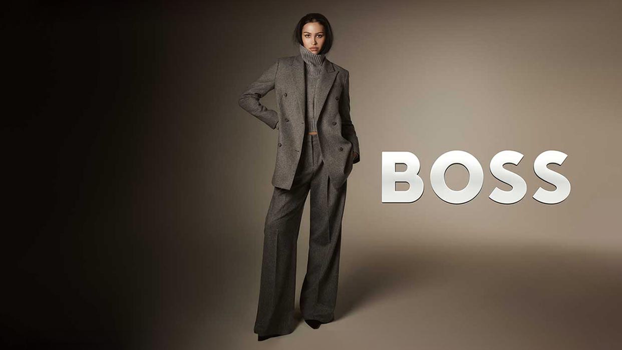 Frau steht mit einer Hand in der Hosentasche, trägt grauen Anzug mit weitem Bein und Blazer, daneben Schriftzug 'BOSS' in Großbuchstaben.