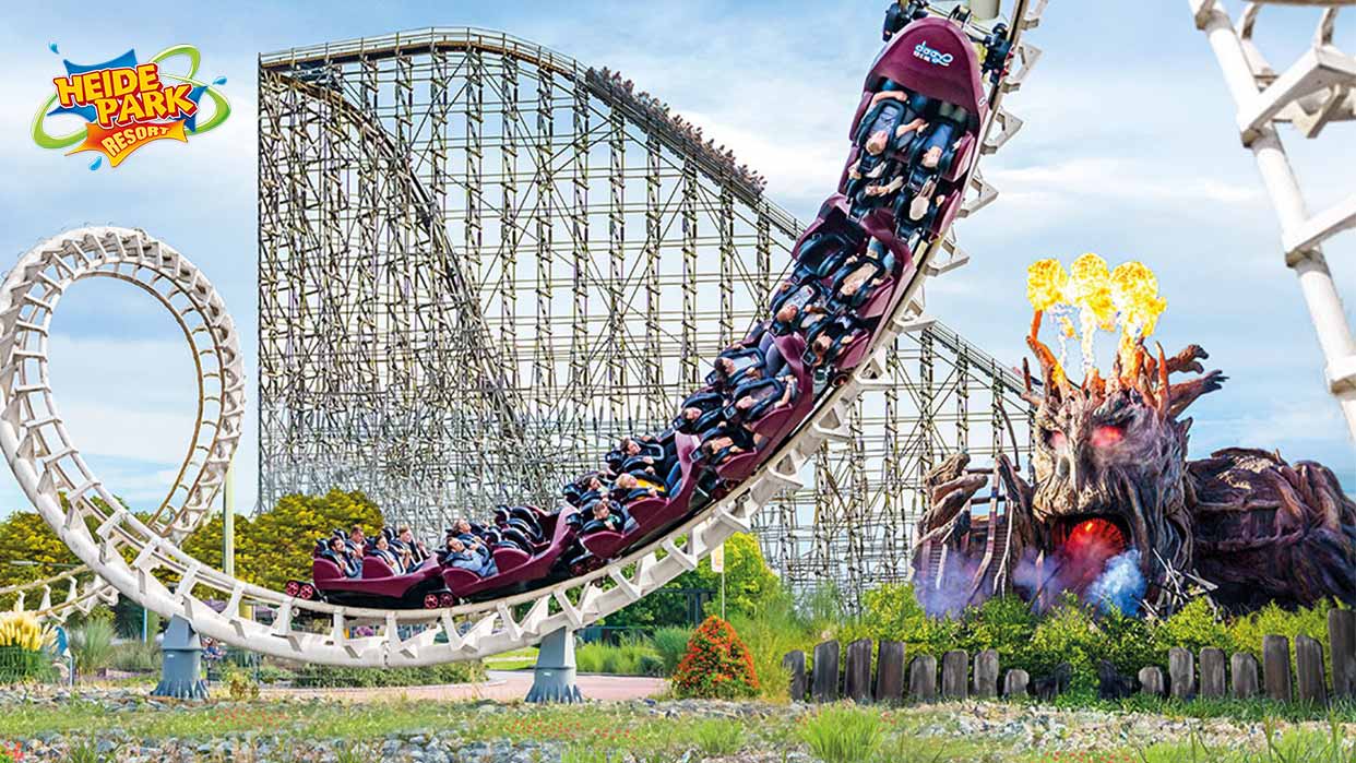 Achterbahn mit Looping und Fahrgästen vor einer großen Holzachterbahn und einer feuerspeienden Baumskulptur mit roten Augen.