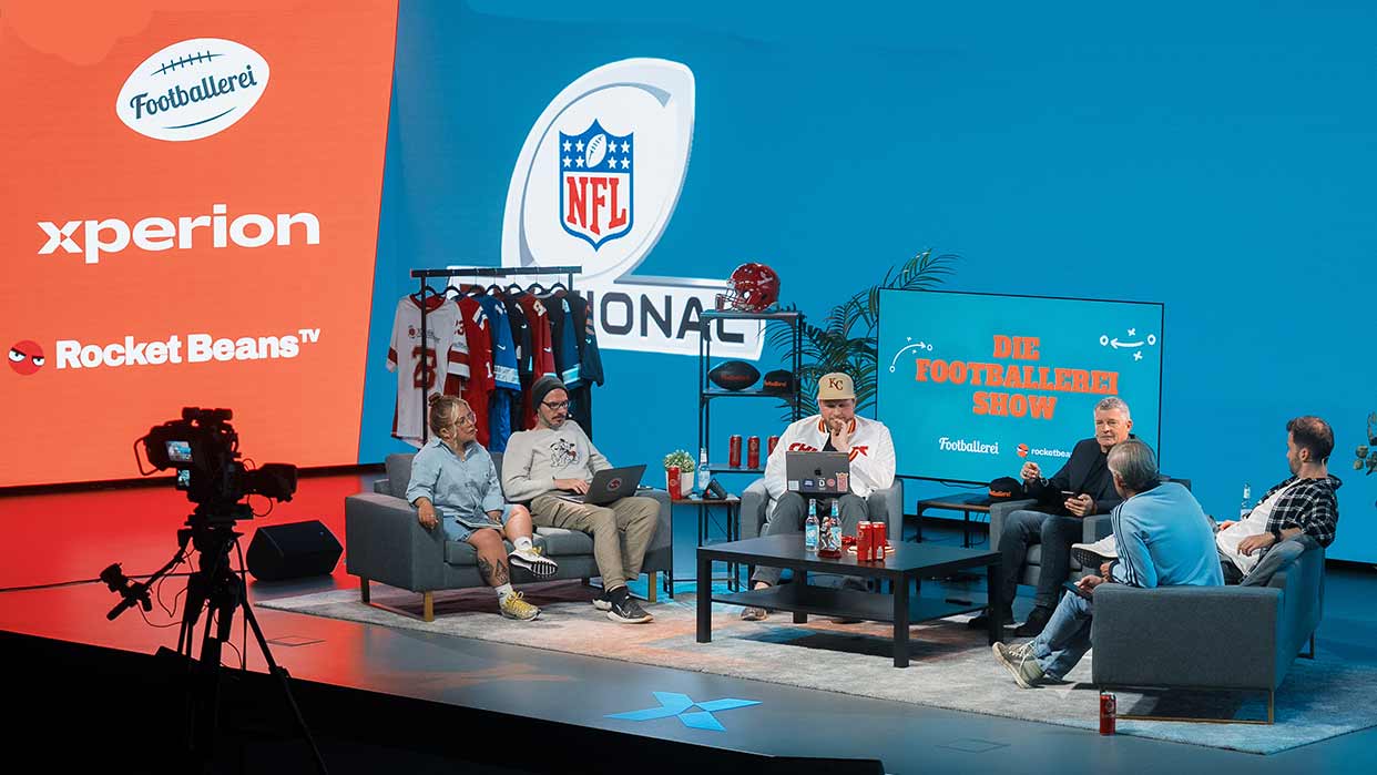 Fünf Personen sitzen in einem Studio mit NFL-Logo, zwei Sofas, einem Fernseher mit Schriftzug 'DIE FOOTBALLEREI SHOW' und rotem Hintergrund mit Logos von xperion und Rocket Beans TV.