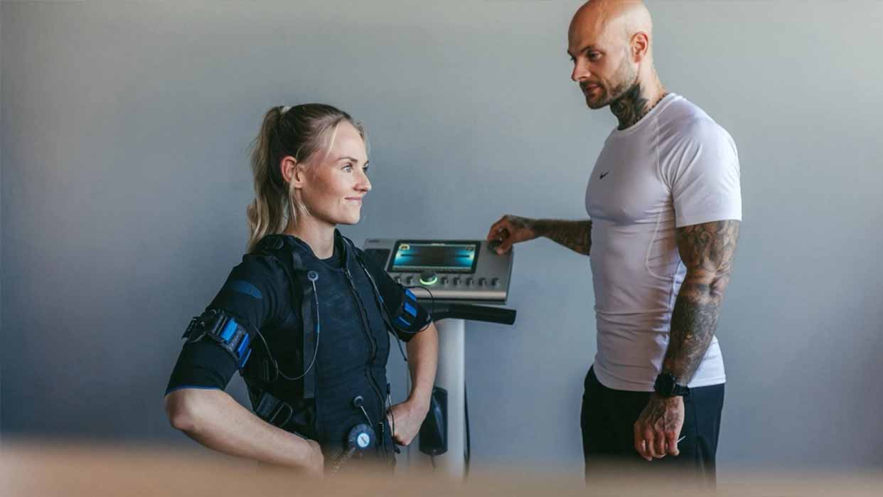 Frau trägt EMS-Anzug und steht mit Händen in der Hüfte vor einer Waage, daneben steht ein tätowierter Mann in Sportkleidung.