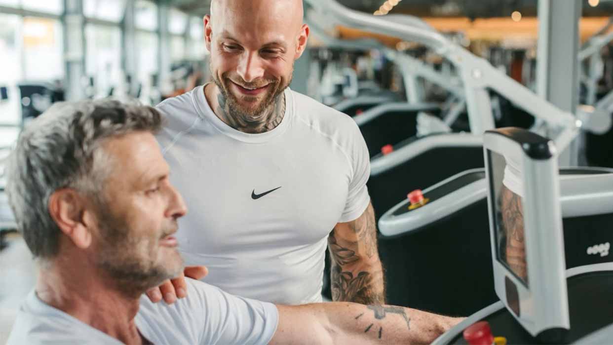 Zwei Männer im Fitnessstudio, einer mit weißem T-Shirt und tätowierten Armen, sitzen an einem Trainingsgerät.
