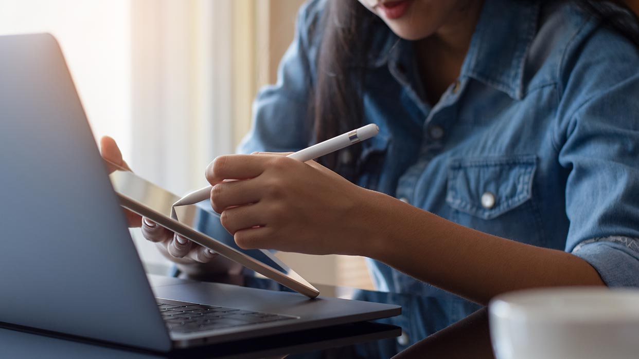 Frau in Jeanshemd nutzt Tablet mit Stift vor offenem Laptop, hält Smartphone in der Hand