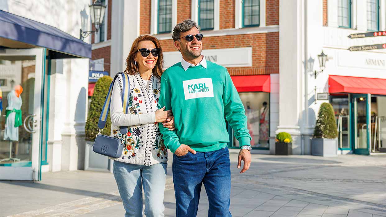 Paar geht Arm in Arm vor einem Designer-Outlet mit Schaufenstern und roten Markisen.