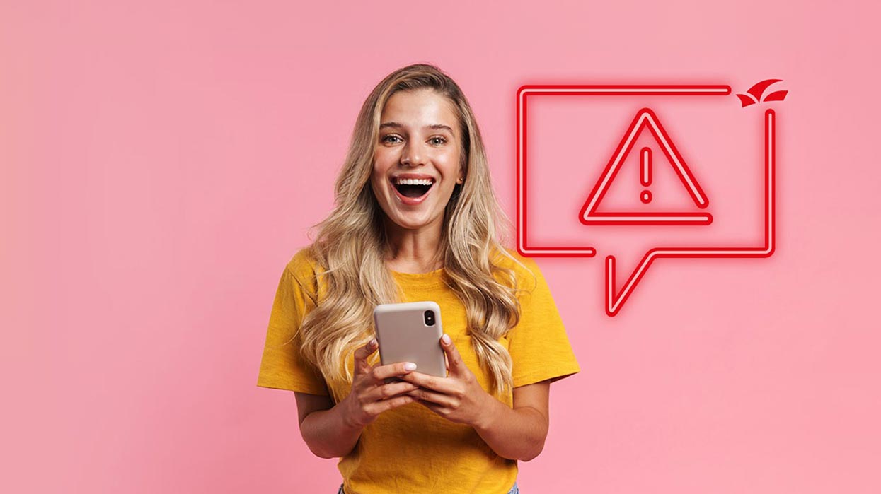 Frau mit langen blonden Haaren in gelbem T-Shirt hält Smartphone vor rosa Hintergrund.