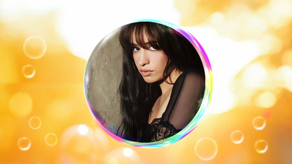Camila Cabello mit langen dunklen Haaren und durchsichtiger schwarzer Bluse umgeben von einer transparenten Seifenblase vor unscharfem, goldenem Hintergrund.