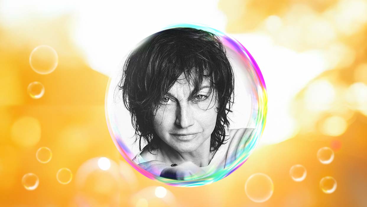 Gianna Nannini mit nassem, dunklem Haar und direktem Blick, umgeben von einer transparenten Seifenblase vor unscharfem orangefarbenem Hintergrund mit Lichtreflexen.