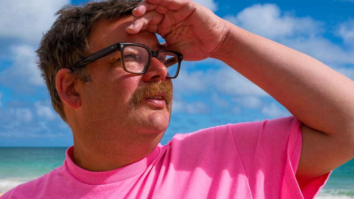 Erobique mit Brille und pinkem T-Shirt hält die Hand über die Augen und blickt zum Himmel am Strand mit Meer und blauem Himmel im Hintergrund.