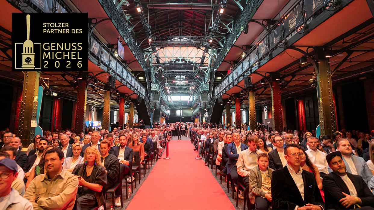 Große Menschenmenge sitzt in einer Halle mit rotem Mittelgang und sichtbarer Stahlkonstruktion im Dachbereich.