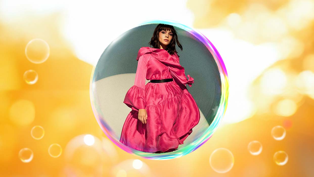 Nora Jones in einem pinkfarbenen, voluminösen Kleid, umgeben von einer transparenten Seifenblase vor einem goldgelben Hintergrund.