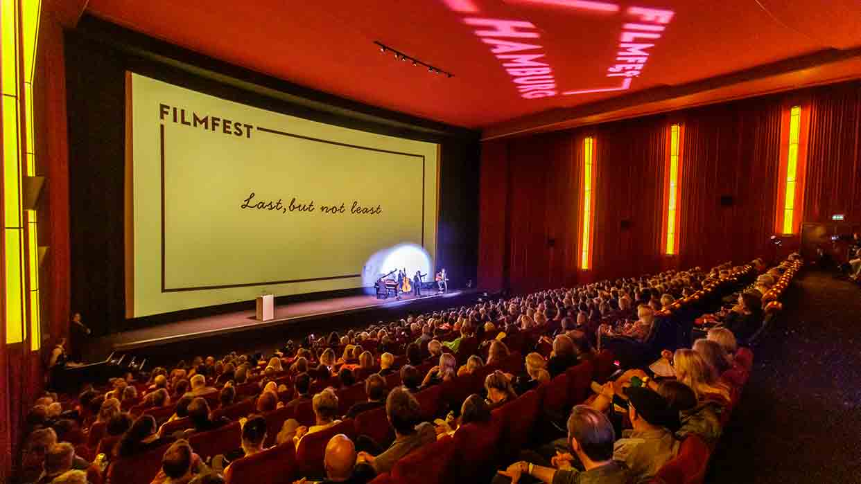 Volles Kino mit Publikum bei Filmfest, auf Leinwand Text 'Last, but not least', Bühne mit vier Personen.