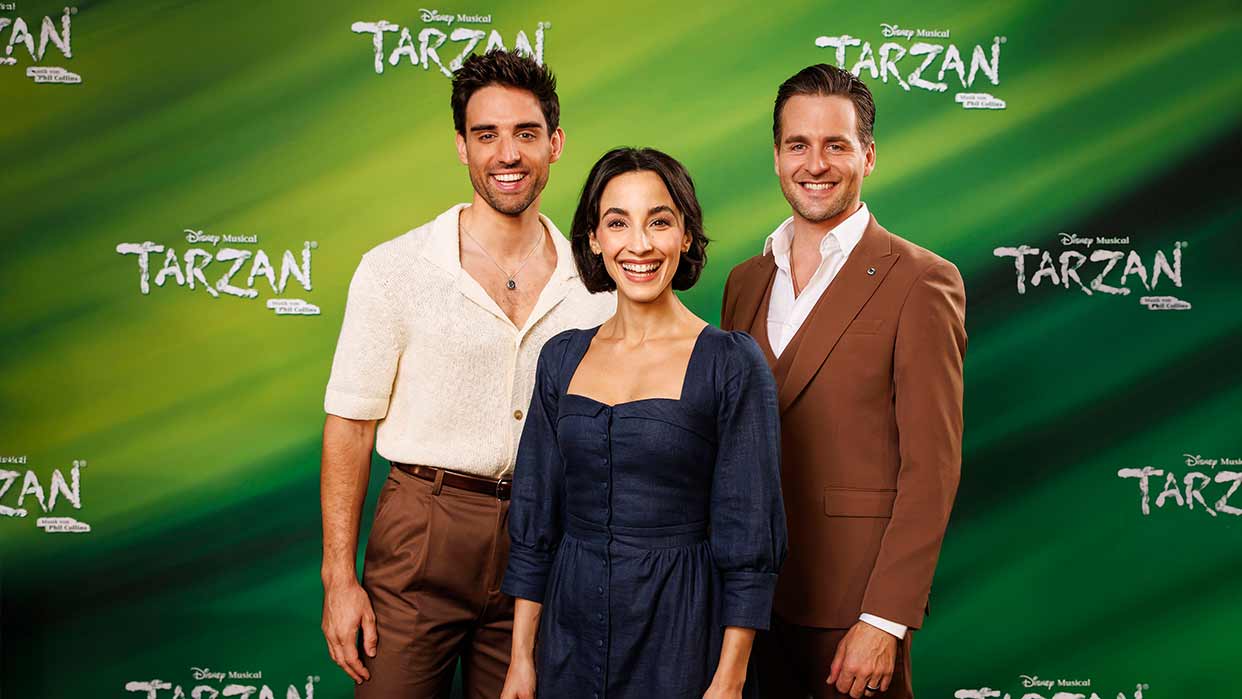 Drei Personen vor grünem Hintergrund mit 'Disney Musical Tarzan' Schriftzug, mittlere Person trägt dunkelblaues Kleid, rechte Person braunen Anzug, linke Person weißes Hemd.