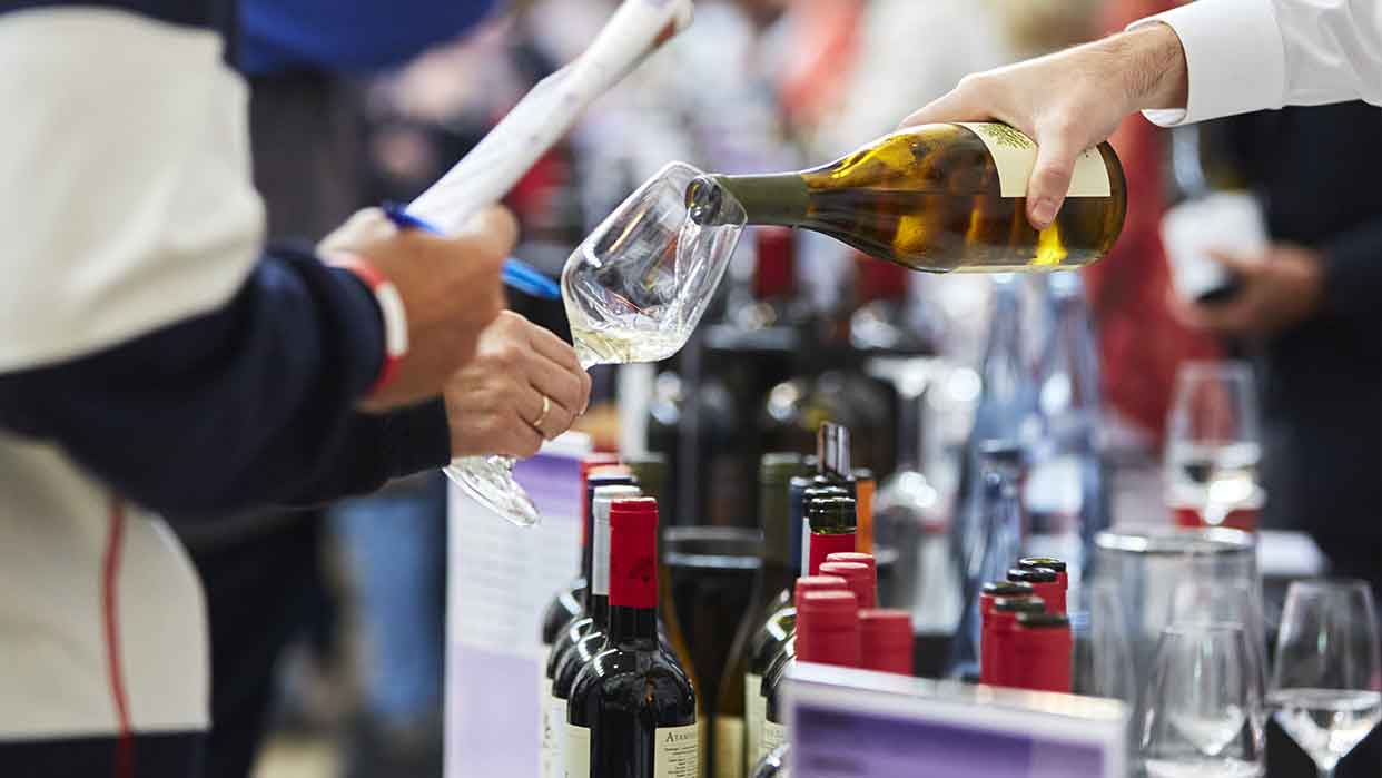 Eine Person gießt Weißwein aus einer Flasche in ein Glas. Im Hintergrund stehen mehrere Weinflaschen mit roten Kappen auf einem Tisch. Die Veranstaltung scheint eine Weinmesse zu sein.