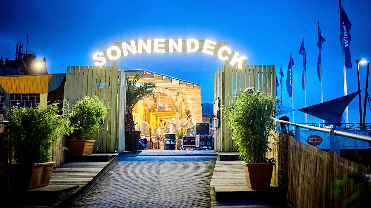 Eingang zu einem beleuchteten Holzdeck mit dem Schriftzug 'SONNENDECK' und großen Pflanzen in Töpfen bei Abendlicht.