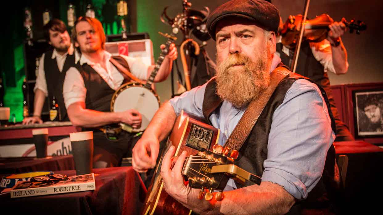 Ein Musiker mit Bart spielt Gitarre, trägt eine Weste und ein Hemd. Im Hintergrund stehen zwei weitere Musiker mit Banjo und Geige. 