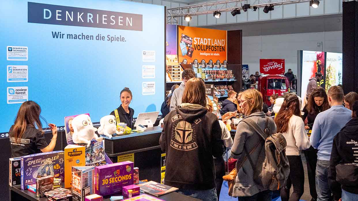 Messestand von DenkRiesen auf der Spielemesse Hamburg. Besucher stehen an einem Tisch mit verschiedenen Brettspielen und Plüschtieren. Im Hintergrund sind weitere Stände sichtbar.
