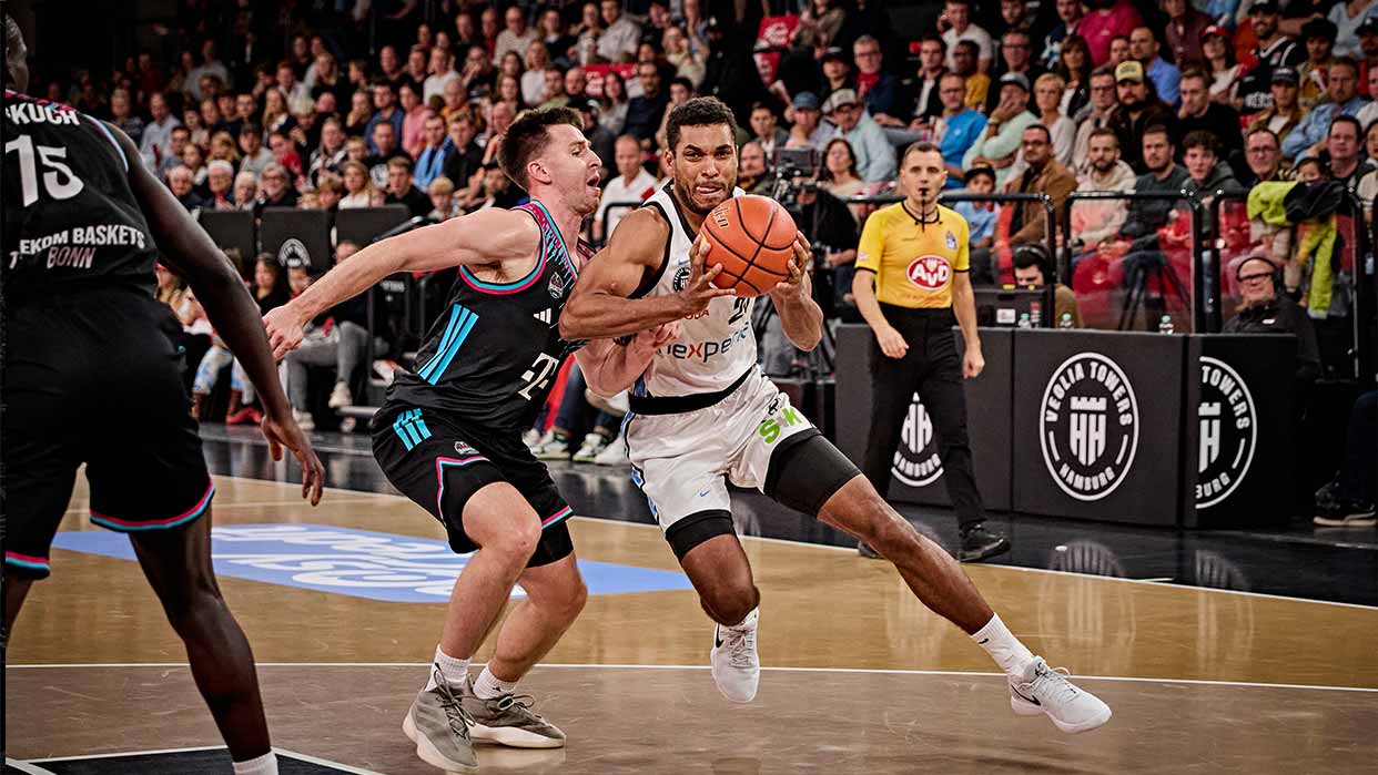 Basketballspieler in weißem Trikot dribbelt den Ball, wird von einem Spieler in schwarzem Trikot verteidigt, im Hintergrund Zuschauer und Schiedsrichter in gelbem Trikot.