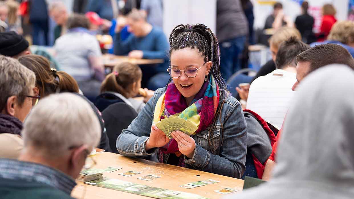 Eine Frau mit geflochtenen Haaren hält eine Handvoll Spielkarten in einem Raum voller Menschen, die an Tischen spielen.