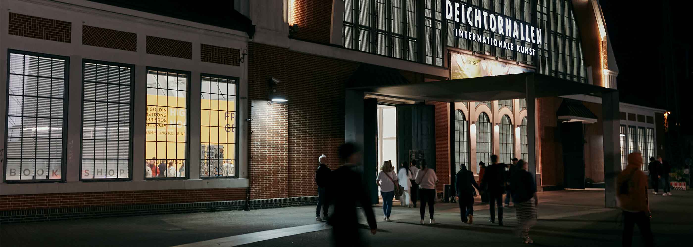 Menschen gehen nachts auf einen beleuchteten Backsteinbau mit dem Schriftzug 'DEICHTORHALLEN INTERNATIONALER KUNST' zu.