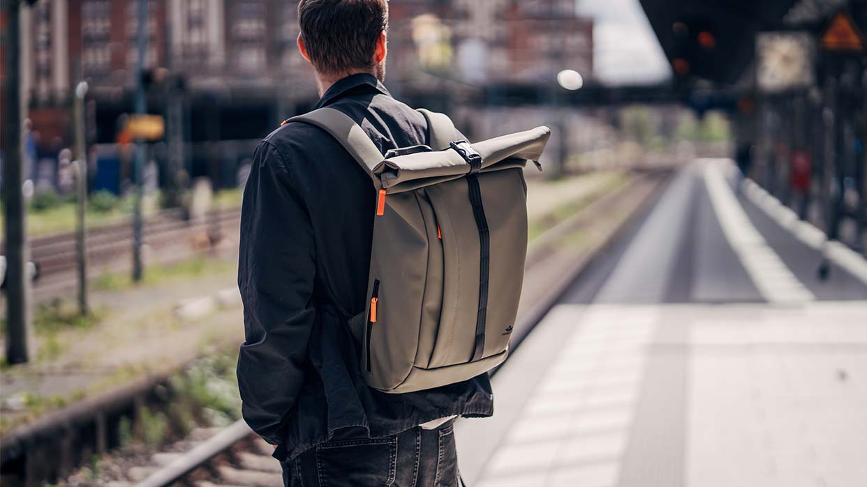 Mann mit grauem Freibeutler Rucksack, steht an einem Bahnsteig. Der Rucksack hat einen Rollverschluss und orangefarbene Akzente.