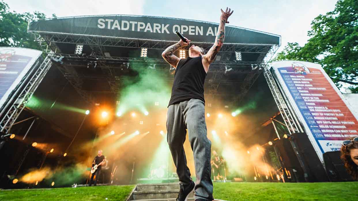 Ein Sänger mit tätowierten Armen trägt ein schwarzes Tanktop und graue Hosen. Er steht auf einer Bühne mit Scheinwerfern und einem Banner 'STADTPARK OPEN AIR' im Hintergrund.