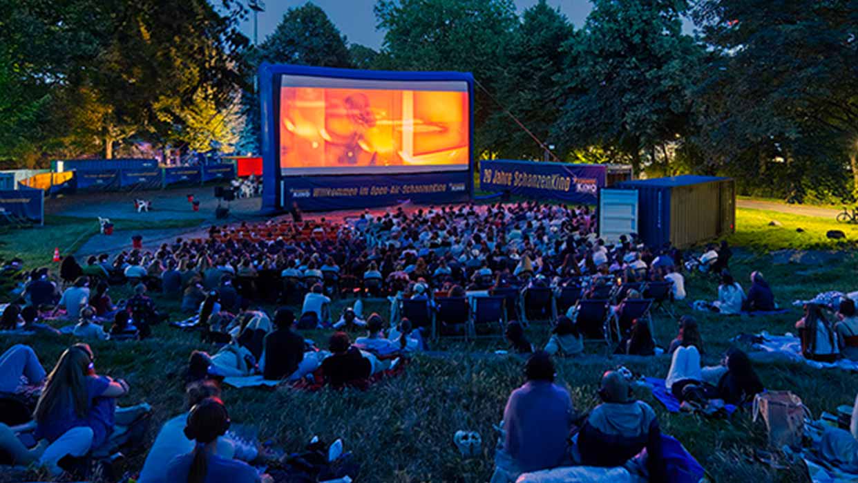 Abendliche Szene im LOTTO Hamburg Open-Air-SchanzenKino mit Publikum auf Wiese und Stühlen vor großer Leinwand im Park.