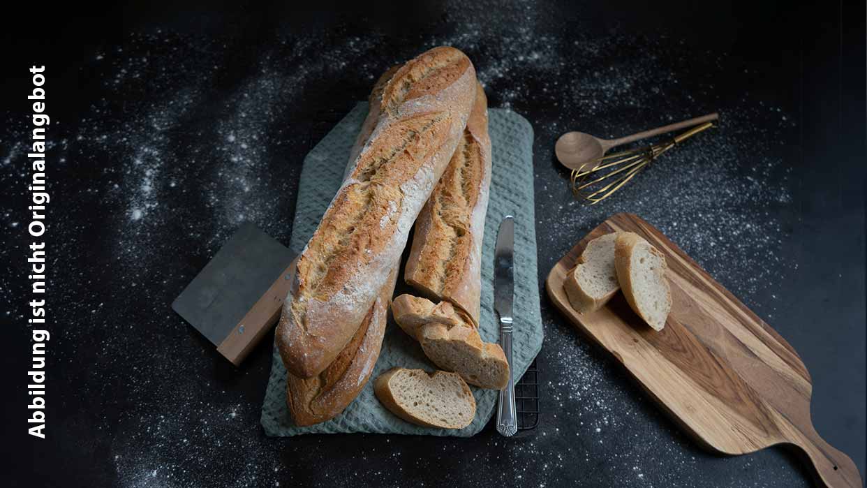 Zwei längliche, rustikale Baguettes auf einem blauen Schneidebrett mit einem Messer, daneben ein Holzbrett mit zwei Brotscheiben und ein Schneebesen, umgeben von Mehlstaub.