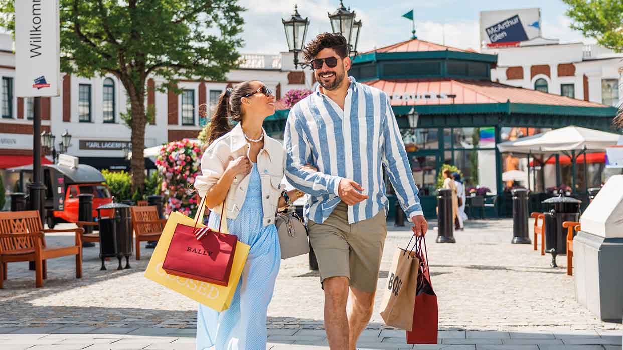 Paar geht mit Einkaufstüten durch das Designer Outlet Neumünster an einem sonnigen Tag.