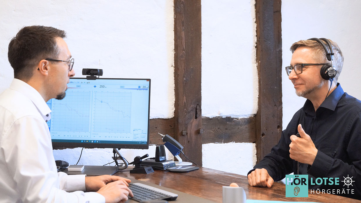 Zwei Männer sitzen an einem Holztisch, einer trägt Kopfhörer und gibt mit Daumen hoch ein Zeichen, der andere bedient eine Tastatur vor einem Monitor mit Diagrammen.