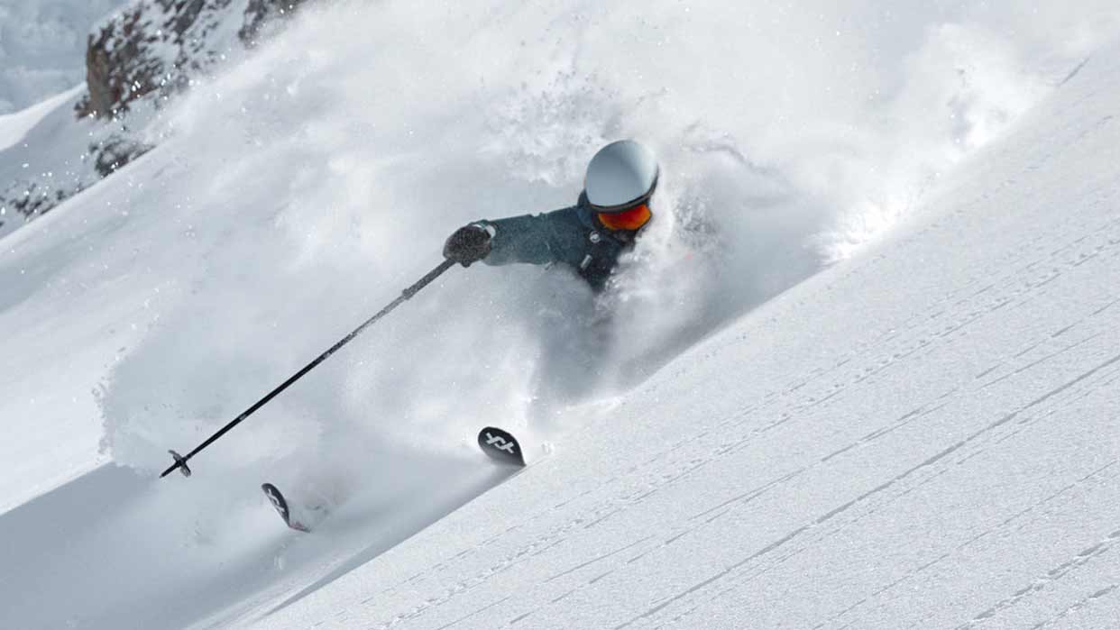 Skifahrer mit Helm und Skibrille fährt durch tiefen Pulverschnee an einem steilen Hang.