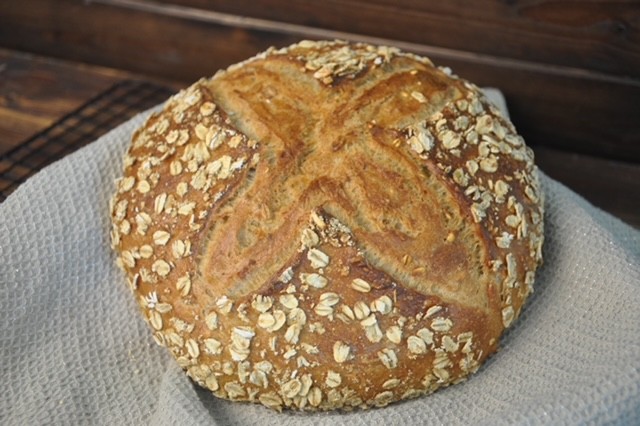 Rundes Vollkornbrot mit Haferflocken bestreut und einem Kreuzschnitt auf der Kruste, auf grauem Tuch liegend.