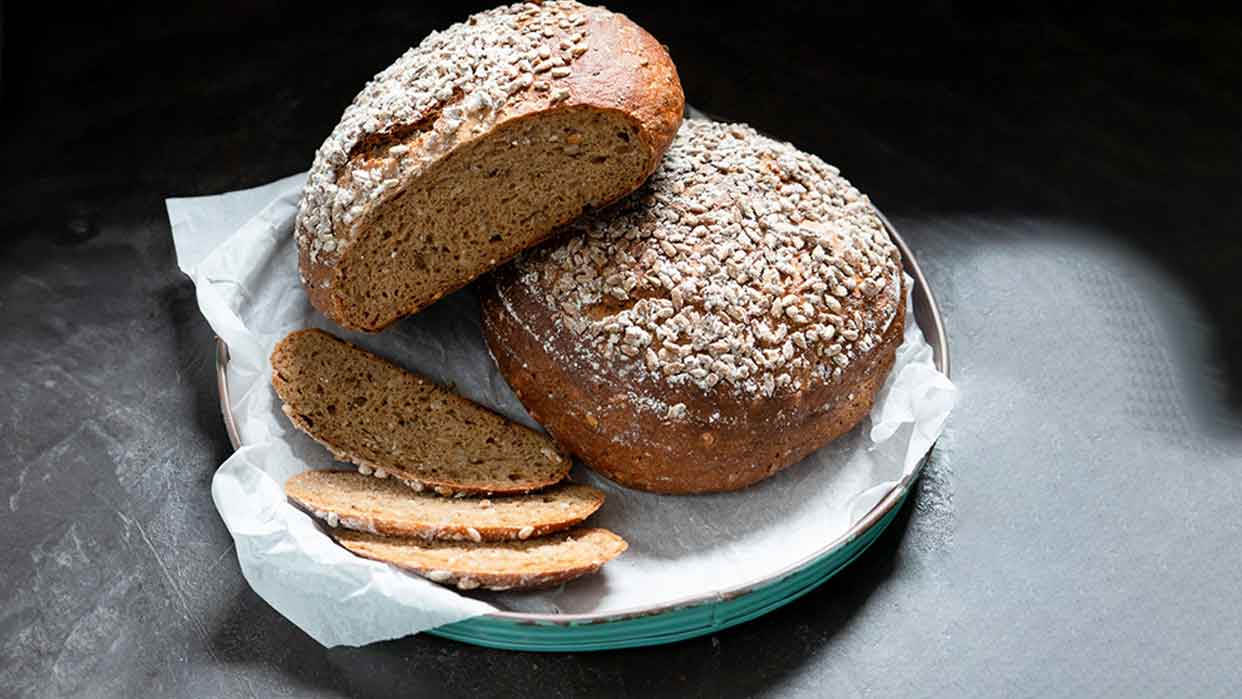 Rundes Brot mit Mehrl und Saaten bestreut auf einem Teller, daneben mehrere Brotscheiben auf Backpapier